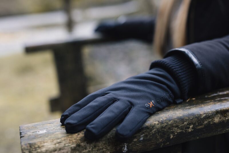 Waterproof-Power-Liner-Gloves-5-3.jpg Close-up of a black gloved hand resting on a weathered wooden bench, showing padded fabric, a small orange logo on the glove and a blurred outdoor background.