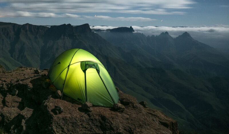 Website1-3.jpg A glowing lime-green tent secured with small rocks on a rocky cliff ledge, overlooking steep, serrated mountain ridges and a sea of low clouds beneath a dim, cloud-streaked sky.