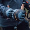 Zenith_Gloves_LS-10-scaled-2.jpg Close-up of gloved hands wearing grey-blue gloves with black leather panels and orange stitching gripping cork-handled trekking poles against a blurred outdoor background.