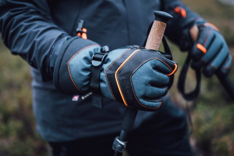 Zenith_Gloves_LS-10-scaled-2.jpg Close-up of gloved hands wearing grey-blue gloves with black leather panels and orange stitching gripping cork-handled trekking poles against a blurred outdoor background.
