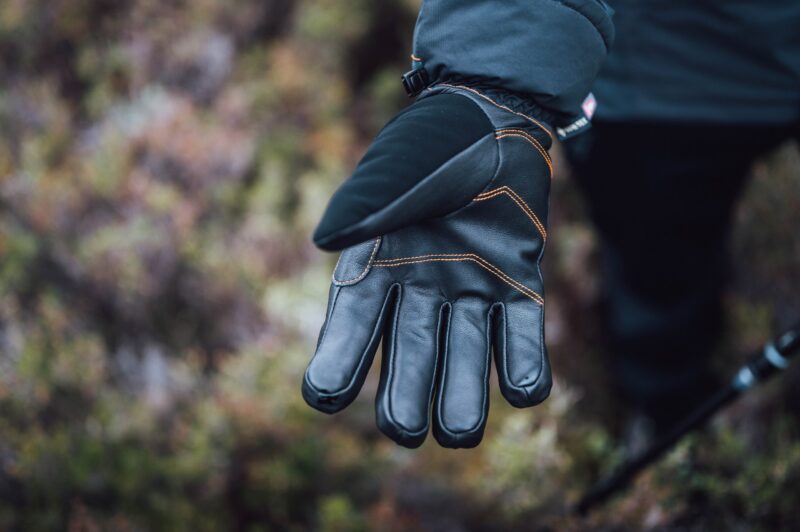 Zenith_Gloves_LS-13-scaled-2.jpg Close-up of an outstretched black insulated glove with leather palm and orange stitching on a person's hand against a blurred natural background.