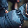 Zenith_Gloves_LS-14-scaled-2.jpg Close-up of a blue and black insulated winter mitten with orange contrast stitching and a black nylon strap and plastic buckle against a blurred outdoor background.