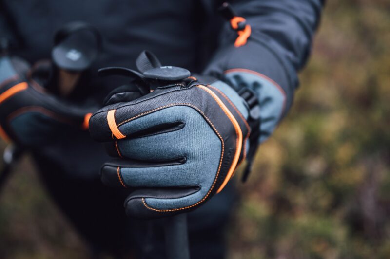 Zenith_Gloves_LS-9-scaled-2.jpg Close-up of a grey and black hiking glove with orange stitching and a wrist strap gripping a trekking pole against a blurred outdoor background.