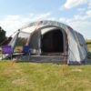 Large grey family tent with an open front and red guy ropes pitched on a grassy field, two folding camping chairs (purple and blue) on the tent porch, with trees to the left and rolling hills under a blue sky with scattered clouds.