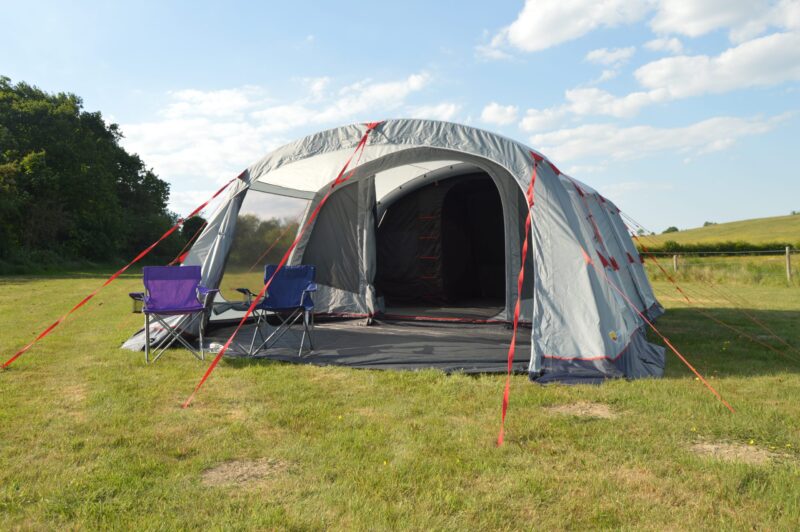 Large grey family tent with an open front and red guy ropes pitched on a grassy field, two folding camping chairs (purple and blue) on the tent porch, with trees to the left and rolling hills under a blue sky with scattered clouds.