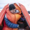 eXtremities-2-scaled-2.jpg Man bundled in an orange sleeping bag and blue down jacket with a black beanie, leaning out of an orange tent to handle a small portable stove while another person lies inside, snowy mountains visible outside.