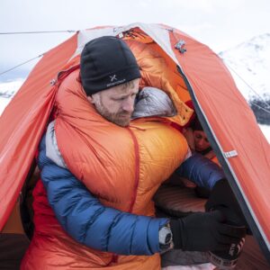 Man bundled in an orange sleeping bag and blue down jacket with a black beanie, leaning out of an orange tent to handle a small portable stove while another person lies inside, snowy mountains visible outside.