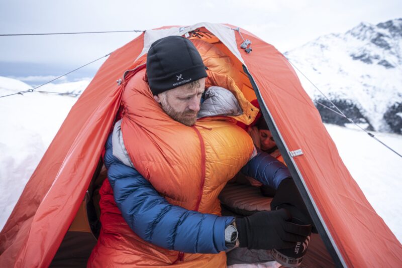 eXtremities-2-scaled-2.jpg Man bundled in an orange sleeping bag and blue down jacket with a black beanie, leaning out of an orange tent to handle a small portable stove while another person lies inside, snowy mountains visible outside.