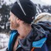 eXtremities-7-scaled-2.jpg Bearded man in profile wearing a black beanie with a reflective strip and a blue hooded jacket, carrying a large backpack against a snowy mountain backdrop.