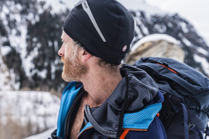 eXtremities-7-scaled-2.jpg Bearded man in profile wearing a black beanie with a reflective strip and a blue hooded jacket, carrying a large backpack against a snowy mountain backdrop.