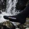 Black performance sock with the 'extremities' logo in white and orange across the instep, resting on wet rocks with a splashing stream or small waterfall in the background