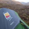Green three-person tent with a grey rainfly showing a Wild Country by Terra Nova TRISAR 3 logo, pitched on mossy upland with rolling hills in the distance.