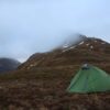 A small green backpacking tent staked on a brown grassy hillside with trekking poles beside it, set against mist‑shrouded, snow‑patched mountain slopes under an overcast sky.