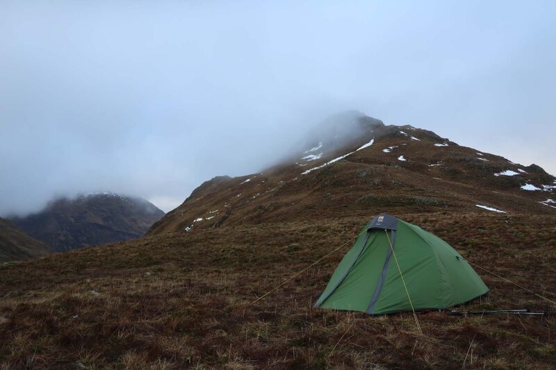 A small green backpacking tent staked on a brown grassy hillside with trekking poles beside it, set against mist‑shrouded, snow‑patched mountain slopes under an overcast sky.