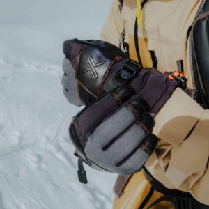 Close-up of insulated grey and black winter gloves with orange stitching and an embossed X logo being adjusted over a tan jacket sleeve against a snowy background.