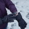 Person in a purple winter jacket uses dark grey insulated gloves to adjust a smartwatch showing colourful activity rings while standing on snow-covered ground.