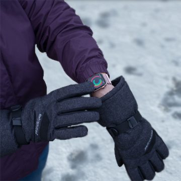 Person in a purple winter jacket uses dark grey insulated gloves to adjust a smartwatch showing colourful activity rings while standing on snow-covered ground.