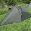 Low-profile olive-green two-person tunnel tent with crossed arched poles and a zippered triangular entrance pitched on a grassy field, another similar tent and bushes visible in the background.