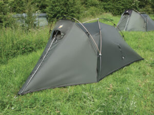 Low-profile olive-green two-person tunnel tent with crossed arched poles and a zippered triangular entrance pitched on a grassy field, another similar tent and bushes visible in the background.