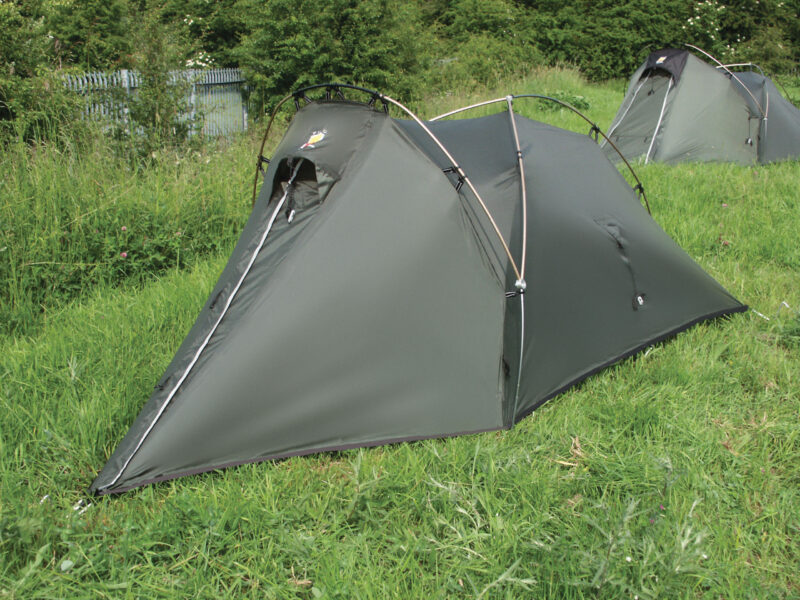 DCF 1.0 Low-profile olive-green two-person tunnel tent with crossed arched poles and a zippered triangular entrance pitched on a grassy field, another similar tent and bushes visible in the background.