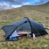 Dark grey single-person tunnel tent pitched on a grassy hillside with the door open to reveal a beige sleeping bag on a red sleeping mat and camping gear (camp stove, sandals and food packets) outside, set against rolling hills and a blue-grey cloudy sky.