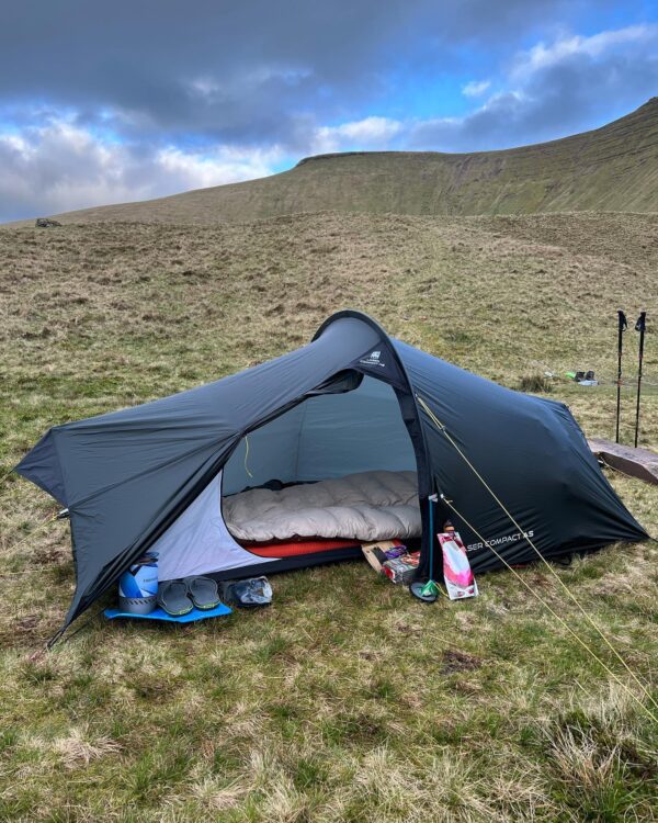 Dark grey single-person tunnel tent pitched on a grassy hillside with the door open to reveal a beige sleeping bag on a red sleeping mat and camping gear (camp stove, sandals and food packets) outside, set against rolling hills and a blue-grey cloudy sky.