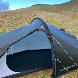 Low-profile dark green Laser Compact A5 tent with its door open to show a pale-blue inner lining and dark groundsheet, staked with yellow guy lines and a blue pole on a grassy upland with rolling hills and a bright blue sky behind.