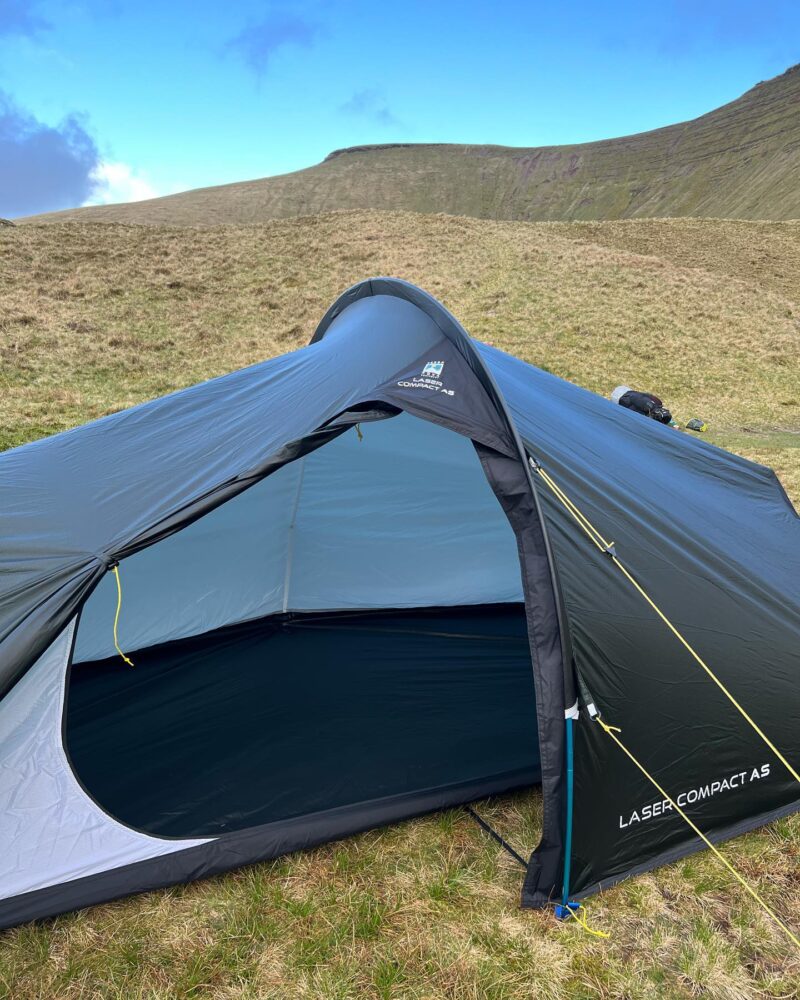 Low-profile dark green Laser Compact A5 tent with its door open to show a pale-blue inner lining and dark groundsheet, staked with yellow guy lines and a blue pole on a grassy upland with rolling hills and a bright blue sky behind.