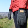terrranova-day-274-scaled-2.jpg Close-up of a person wearing a red quilted jacket and grey knitted gloves with a tan "extremities" label, standing on a grassy country path beside a wooden fence and distant hills under a blue sky.