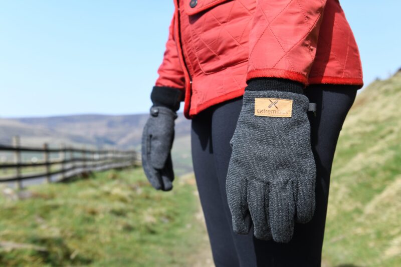 terrranova-day-274-scaled-2.jpg Close-up of a person wearing a red quilted jacket and grey knitted gloves with a tan "extremities" label, standing on a grassy country path beside a wooden fence and distant hills under a blue sky.