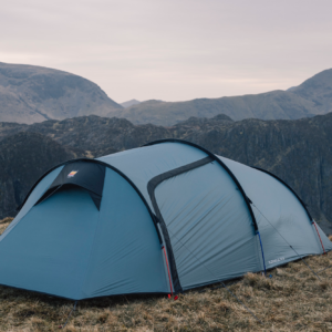Blue tunnel-style camping tent staked on a grassy mountain ridge with guy lines and coloured pole ends, set against layered rocky hills under an overcast sky.