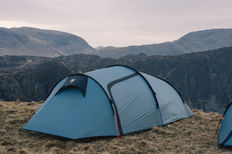 Blue tunnel-style camping tent staked on a grassy mountain ridge with guy lines and coloured pole ends, set against layered rocky hills under an overcast sky.