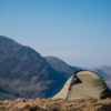 Khaki-green dome tent pitched on a windswept grassy ridge with layered rocky mountains and a clear blue sky in the background.