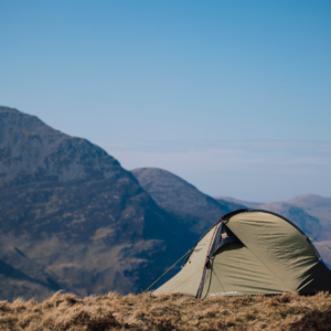 Khaki-green dome tent pitched on a windswept grassy ridge with layered rocky mountains and a clear blue sky in the background.