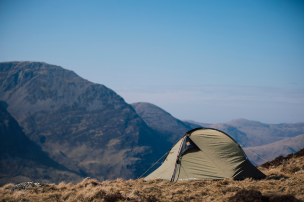 Khaki-green dome tent pitched on a windswept grassy ridge with layered rocky mountains and a clear blue sky in the background.
