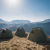 Three olive-green dome tents pitched on a sunlit grassy ridge overlooking layered blue-grey mountains beneath a clear pale-blue sky.