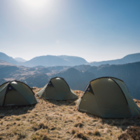Three olive-green dome tents pitched on a sunlit grassy ridge overlooking layered blue-grey mountains beneath a clear pale-blue sky.