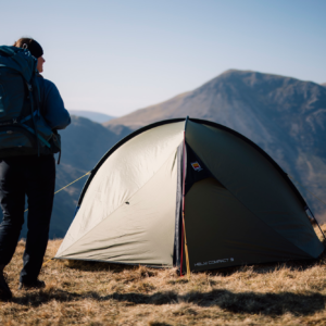 Hiker carrying a large blue backpack stands beside a khaki-green dome tent pitched on a grassy mountain ridge, with sunlit rugged peaks and a clear pale-blue sky in the background.