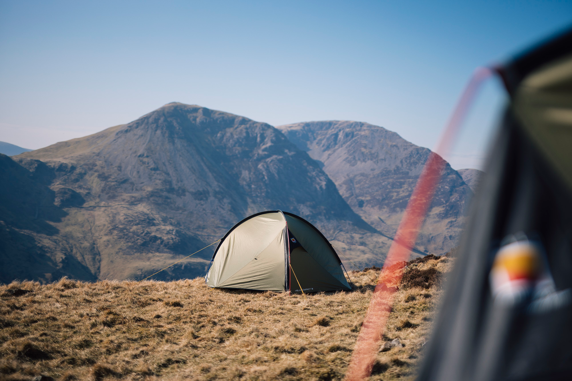 A small green dome tent pitched on a windswept grassy ridge with steep rocky mountains under a clear blue sky, a blurred tent edge and red guyline visible in the foreground.