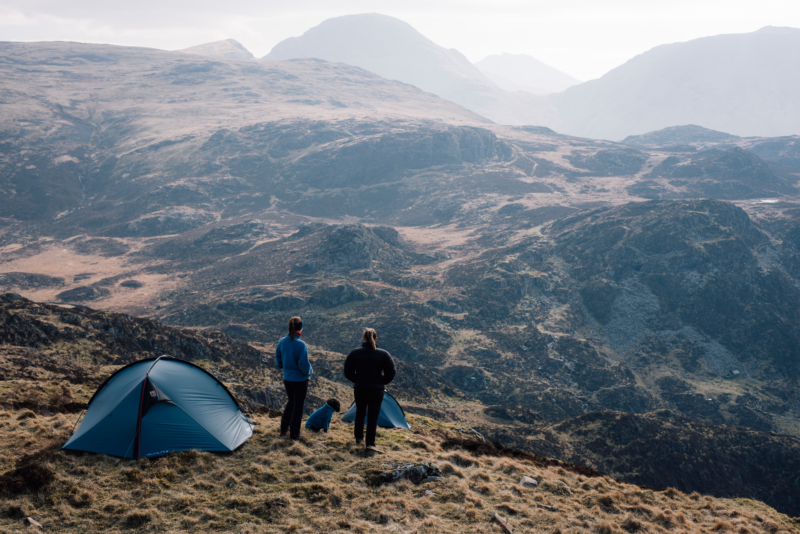 tn_helm_elite_3__ls_2-3.webp Two people and a dog stand beside blue tents on a grassy mountain ridge, looking out over layered rocky hills and misty peaks under a pale sky.