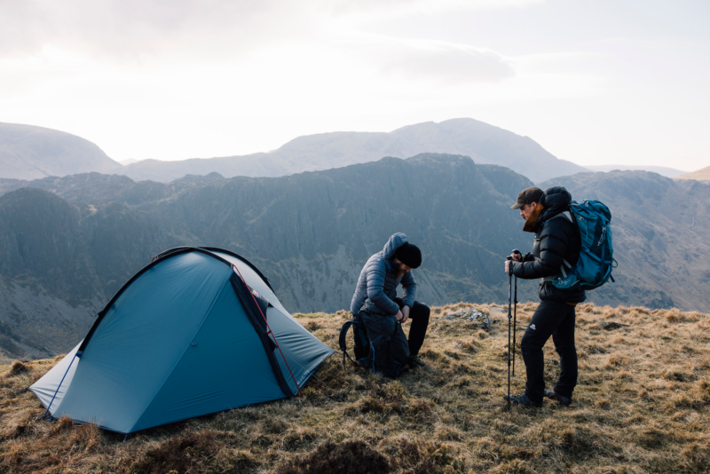 tn_helm_elite_3__ls_4-3.webp Two hikers beside a blue dome tent on a grassy mountain ridge; one sits adjusting a backpack while the other stands with trekking poles and a large rucksack against a backdrop of layered craggy peaks.