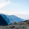 tn_solar_horizon_2__ls5-3.webp Dark blue camping tent pitched on a windswept grassy hillside with layered blue mountains receding under a pale, cloud-streaked sky.