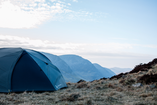 tn_solar_horizon_2__ls5-3.webp Dark blue camping tent pitched on a windswept grassy hillside with layered blue mountains receding under a pale, cloud-streaked sky.