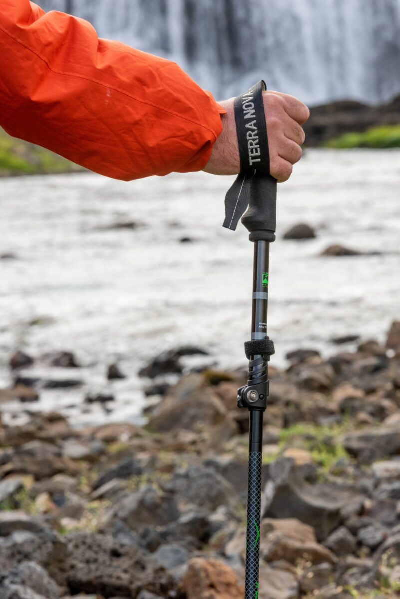 Hand in an orange jacket gripping a trekking pole with a Terra Nova wrist strap, set against a rocky riverbank and a waterfall in the background