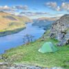 Green two-person tent pitched on a grassy ledge beside rocky crags, overlooking a long blue lake winding between sunlit hills and a partly cloudy sky.