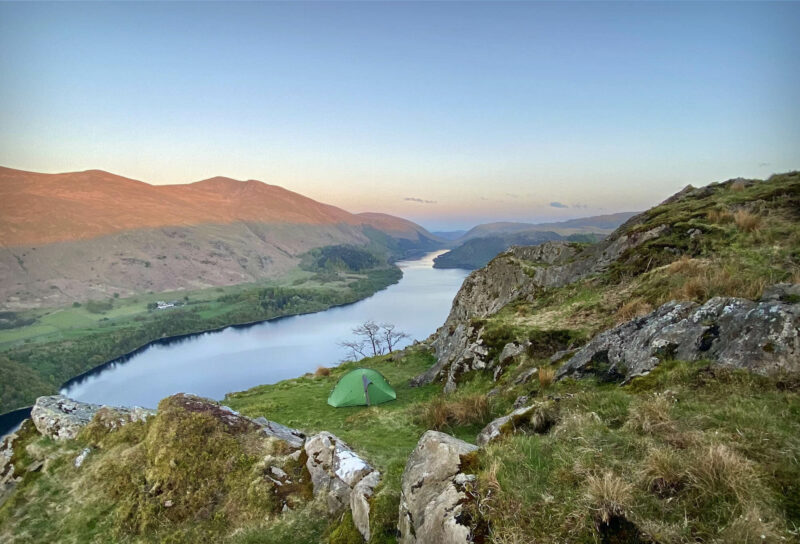 Green tent pitched on a grassy ledge among rocky outcrops, overlooking a long, narrow lake winding between sunlit hills and rugged slopes beneath a pale blue sky at dusk.