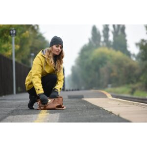 Woman in a yellow raincoat and grey beanie crouches on a misty train platform, reaching into a brown leather handbag by the yellow safety line with tracks and trees fading into the distance.