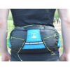 Person's lower back wearing a blue-and-black waist pack with neon green bungee cords and a small white mountain logo, photographed outdoors on a path with blurred walkers in the background.
