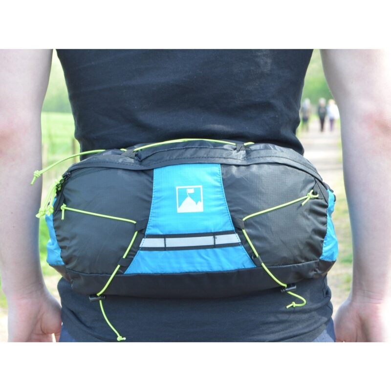 Person's lower back wearing a blue-and-black waist pack with neon green bungee cords and a small white mountain logo, photographed outdoors on a path with blurred walkers in the background.