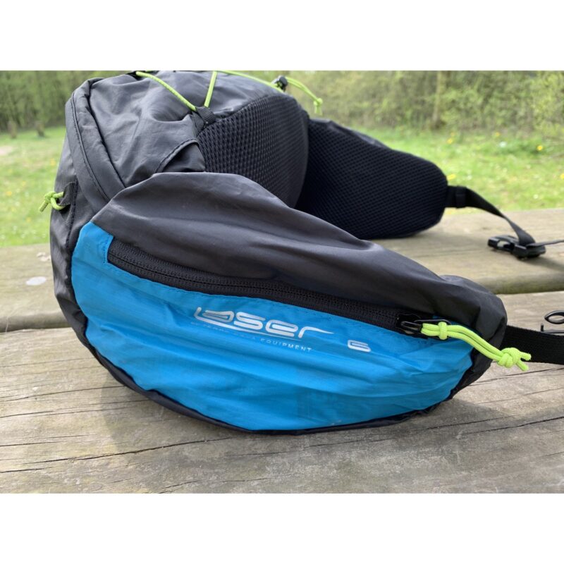 Blue and dark grey waist pack with neon green zipper cords and a white "oser 6" logo on the front, showing a padded mesh back, resting on a weathered wooden picnic bench with grass and trees in the background.